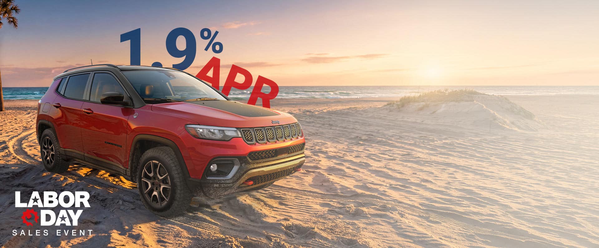 A red 2026 Jeep Compass Trailhawk with black hood insert, parked on a sandy beach at sunset. 1.9% APR. Labor Day Sales Event.
