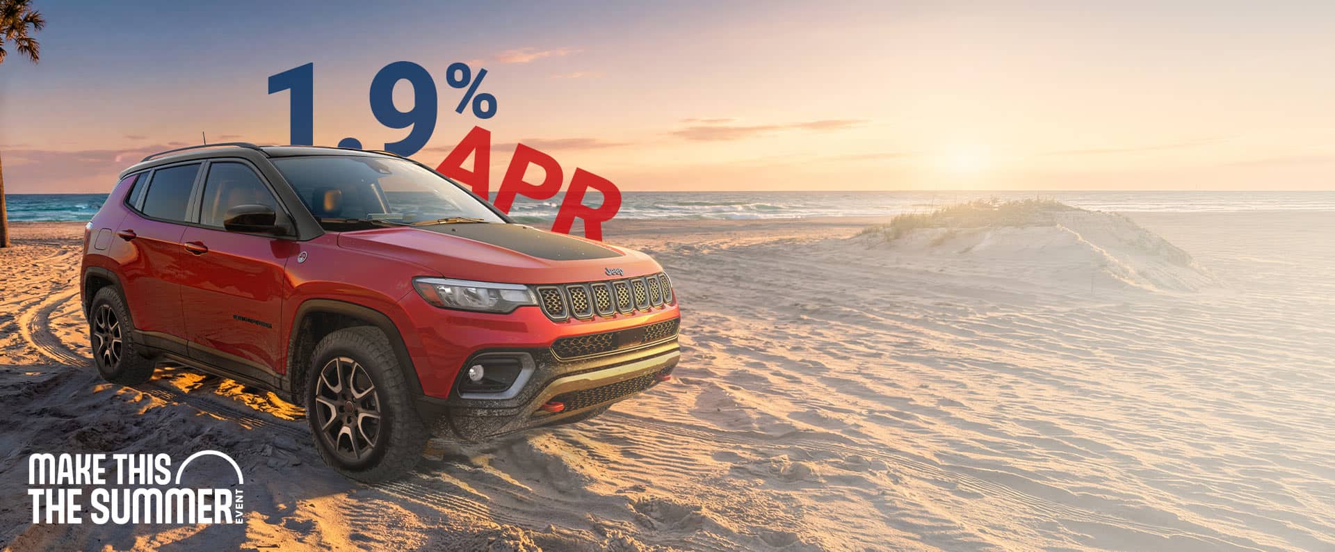 A red 2026 Jeep Compass Trailhawk with black hood insert, parked on a sandy beach at sunset. 1.9% APR. Make This The Summer Event.