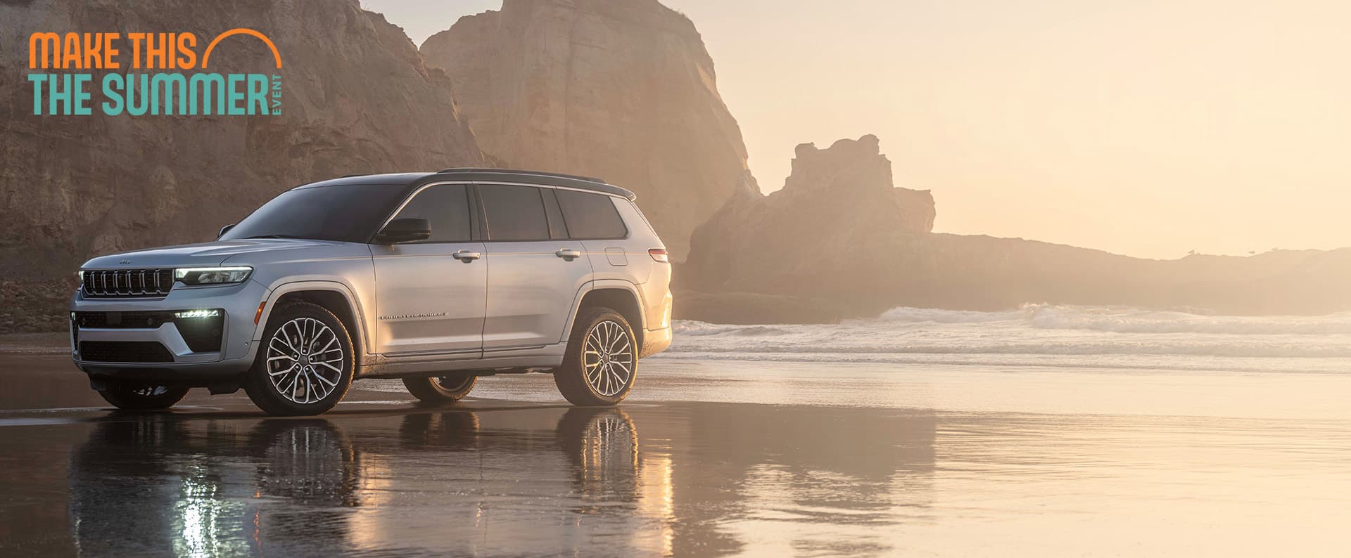 A silver 2026 Jeep Grand Cherokee Summit, parked on a wet beach with rock formations in the background. Make This The Summer Event.