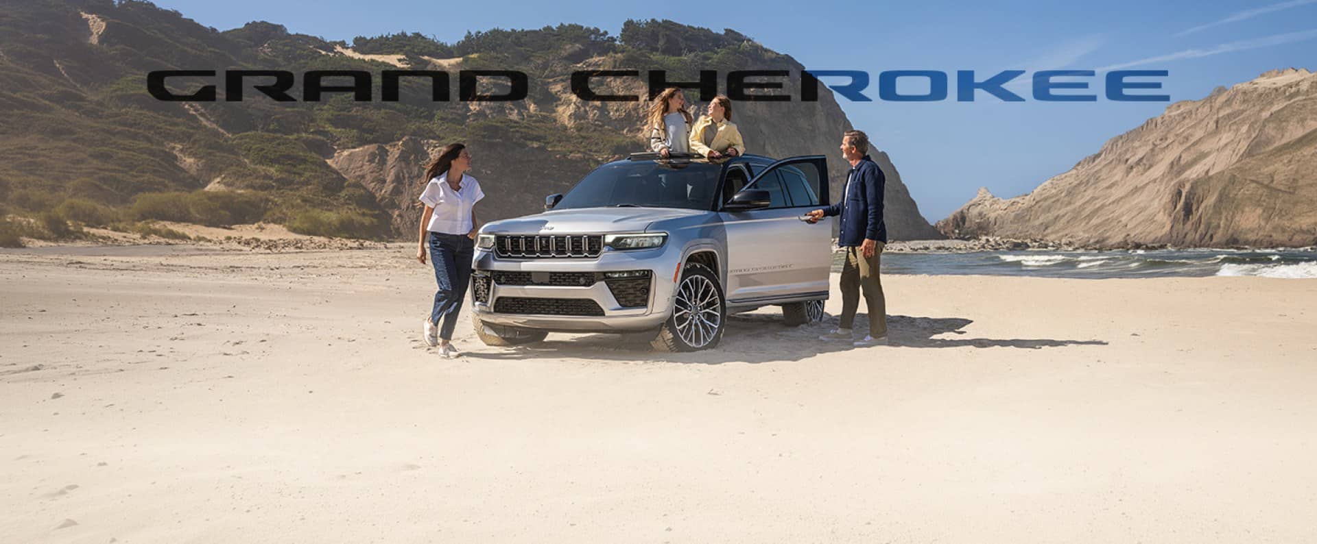 A silver Jeep Grand Cherokee Summit sits parked on the beach surrounded by grassy hills, with a family emerging from the vehicle doors and sunroof. The words 'GRAND CHEROKEE' are superimposed behind it.