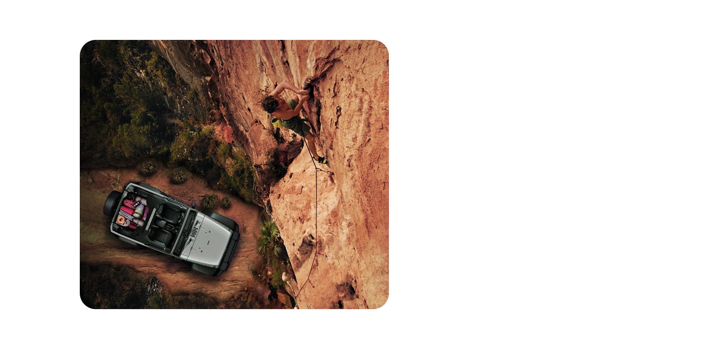 An overhead angle of a silver 2024 Jeep Wrangler with its roof removed and its cargo area filled with camping gear, parked beside a stone mountain, with a climber scaling the rock wall.