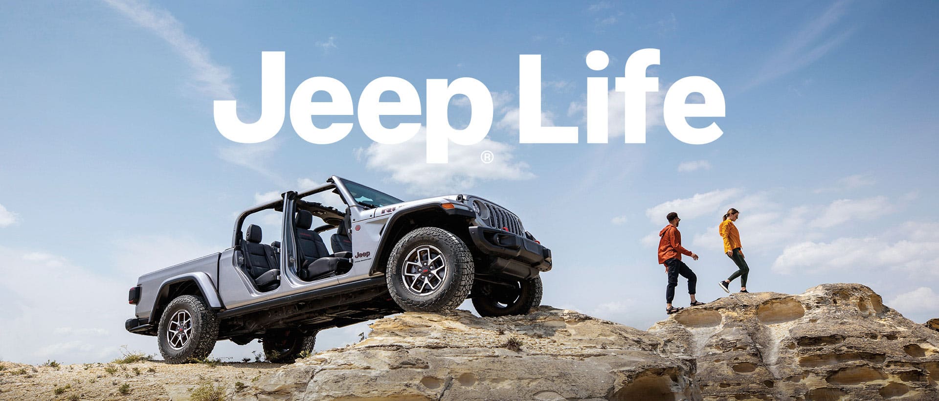 A silver 2024 Jeep Wrangler Rubicon X with its doors and roof removed, parked on an uneven stone surface with its front wheels extended much higher than its rear wheels. Nearby a couple is hiking over rocky outcroppings. Jeep Life.