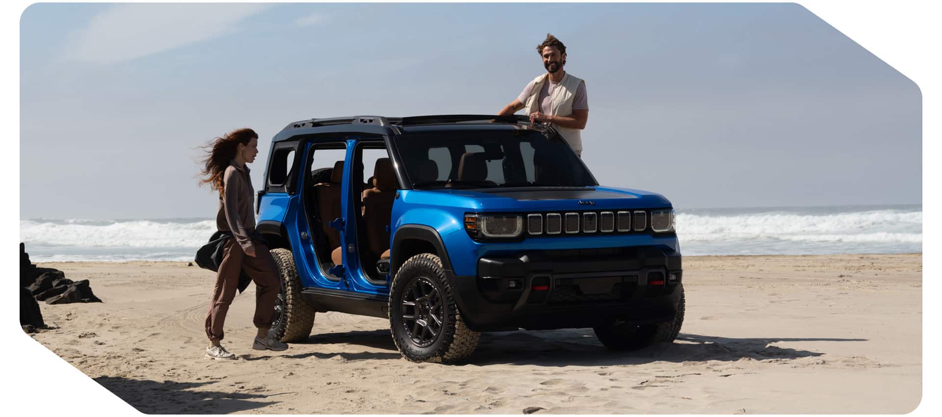 The passenger-side front angle of a blue 2026 Jeep Recon with its doors removed, parked on a sandy beach with a woman and man on each side of the SUV.
