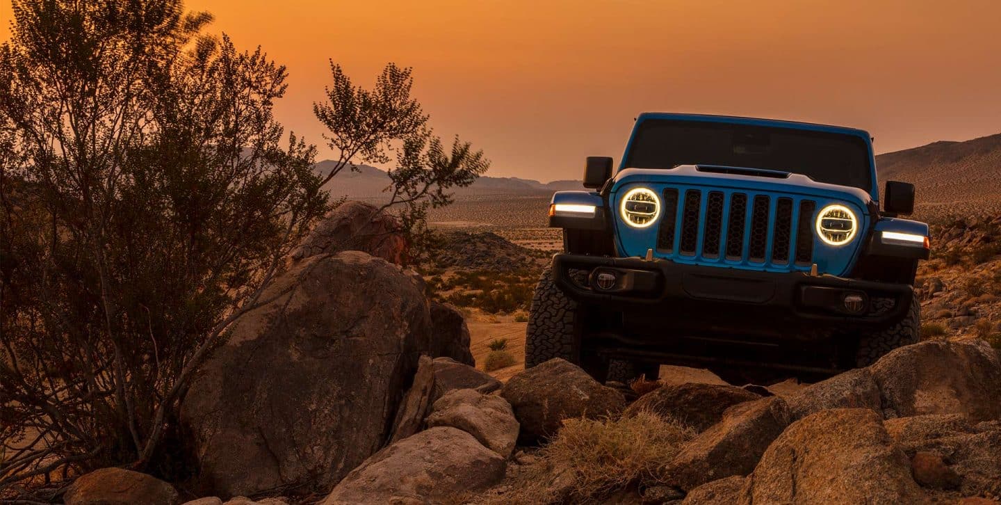 A head-on view of a 2022 Jeep Wrangler Rubicon 392 with its headlamps on, crawling over boulders with a bright orange sky at dusk in the background.