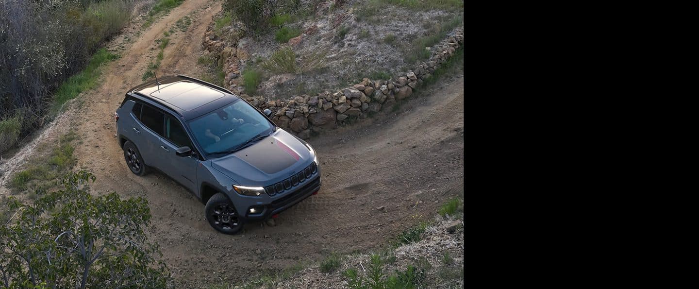 Un Jeep Compass Trailhawk din 2024, condus pe curba strânsă a unui deal de pe un drum de pământ, lângă un gard de piatră.