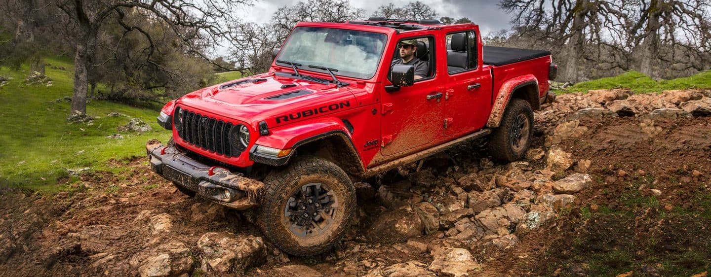 Una Jeep Gladiator Rubicon 2024 roja con el techo desmontado, circulando todoterreno por un sendero rocoso y fangoso.