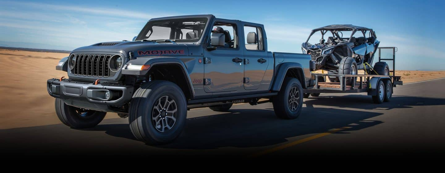 Un Jeep Gladiator Mojave gri 2024 este condus pe o autostradă în timp ce remorchează un ATV pe o remorcă utilitară.