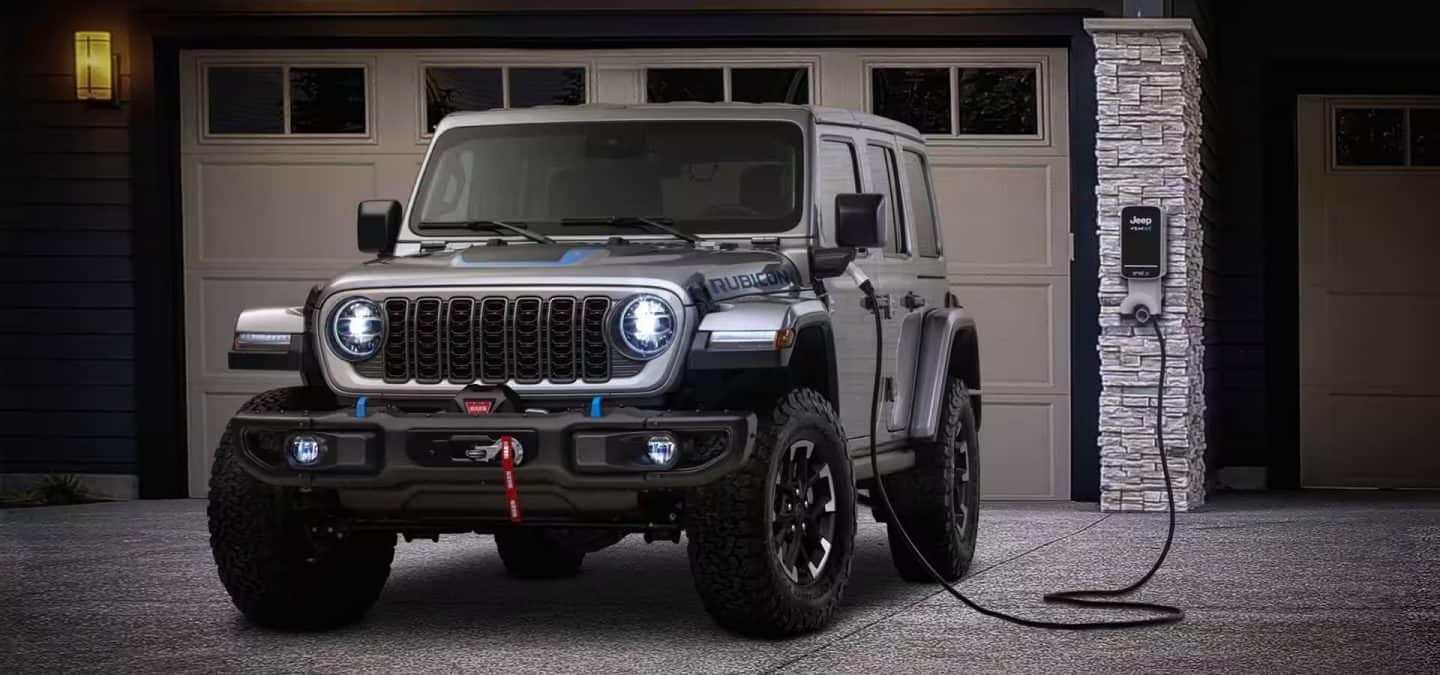 Un Jeep Wrangler Rubicon 4xe estacionado en un garaje con una estación de carga en la pared y el cable de carga enchufado al puerto de carga del vehículo.