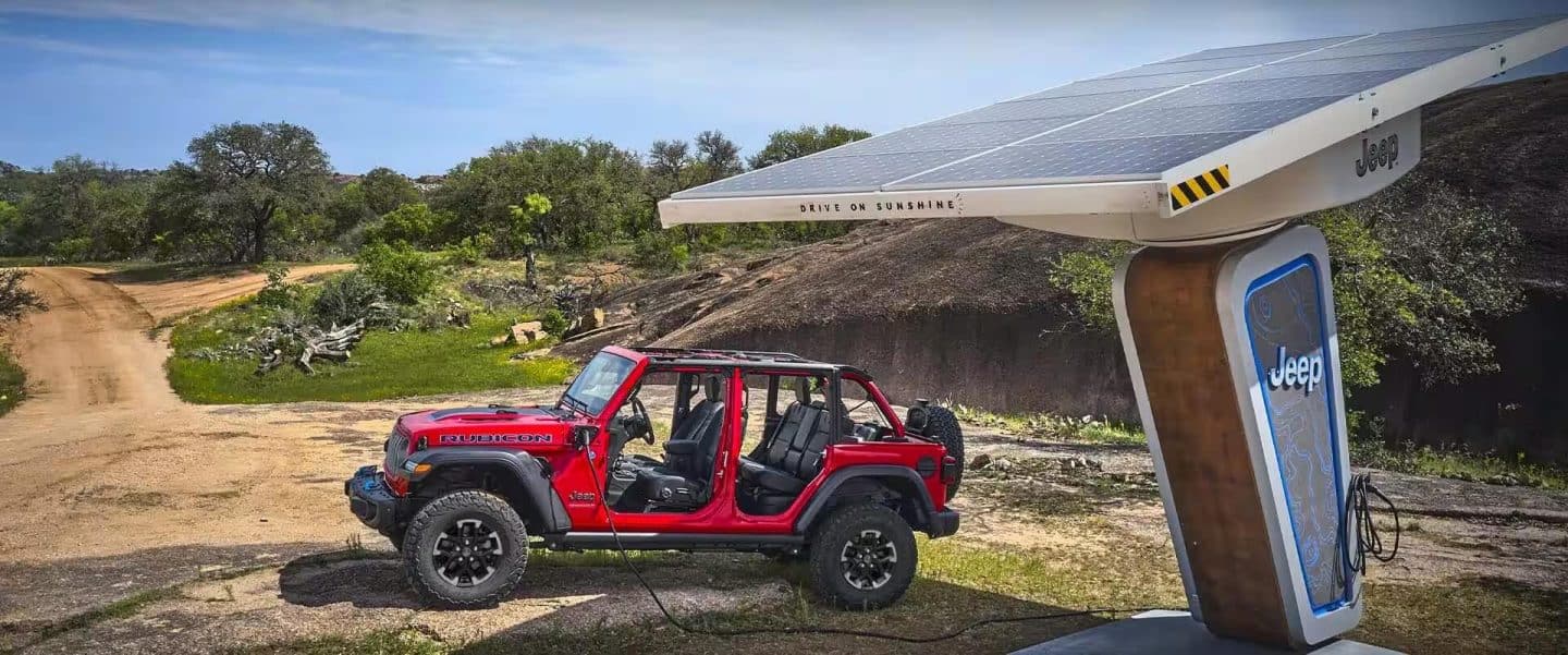 Un Jeep Wrangler Rubicon 4xe 2024 rojo, con las puertas y el techo desmontados, se muestra estacionado junto a una estación de carga con alimentación solar de la marca Jeep. El cable de carga está enchufado al puerto de carga del vehículo.