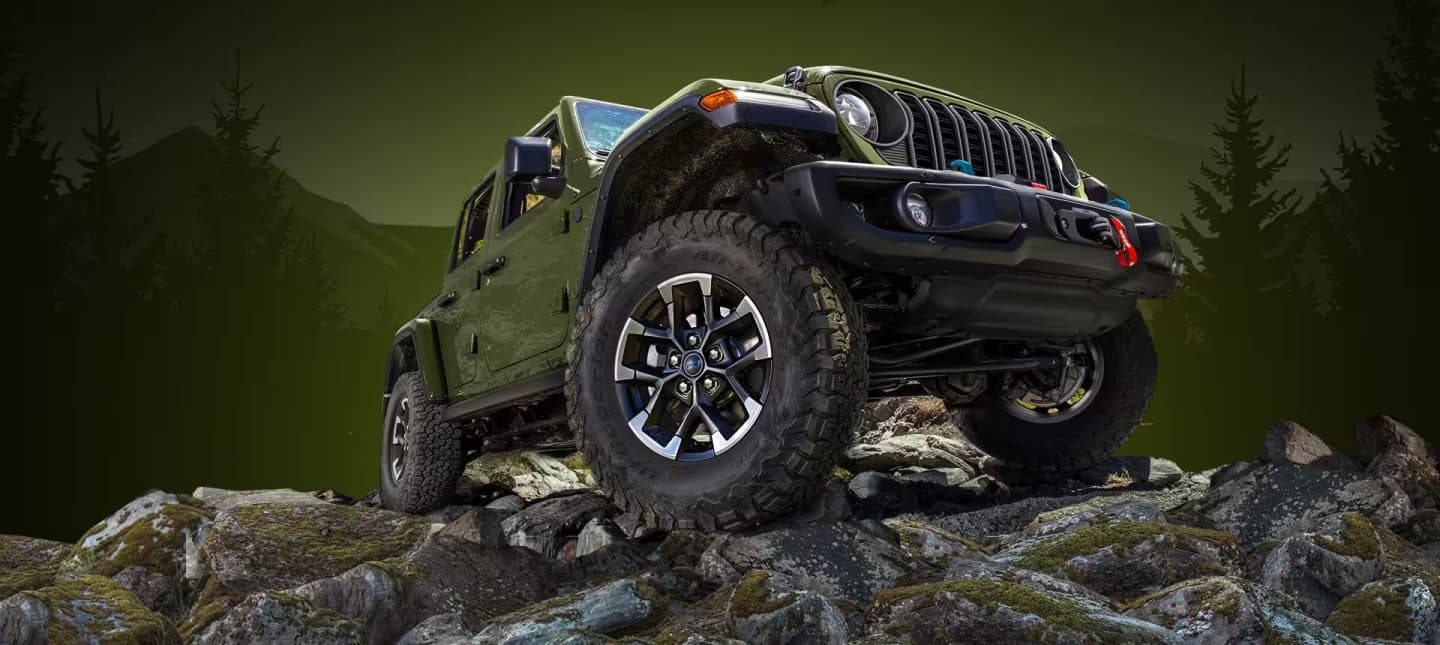 A angle from below of a green 2024 Jeep Wrangler Rubicon 4xe crawling over a stoney mound off-road.