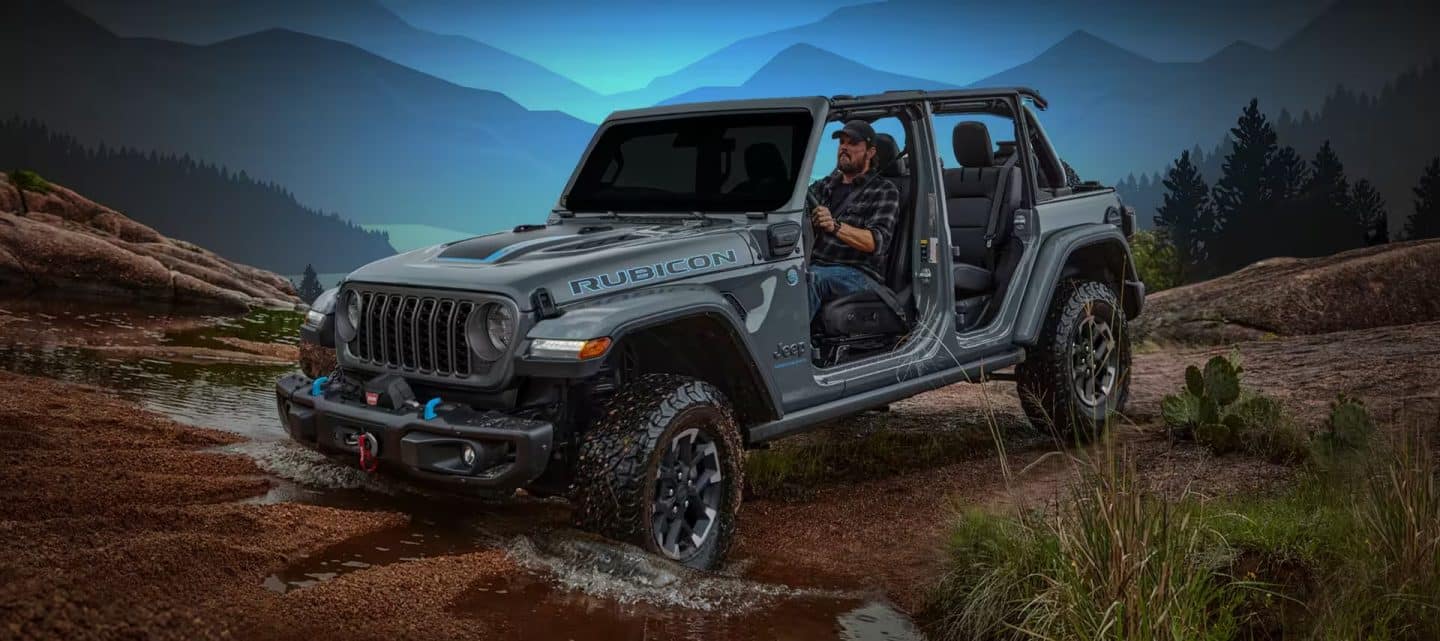 Un Jeep Wrangler Rubicon 4xe 2024 plateado sin las puertas desciende una colina mientras atraviesa un arroyo en las montañas.