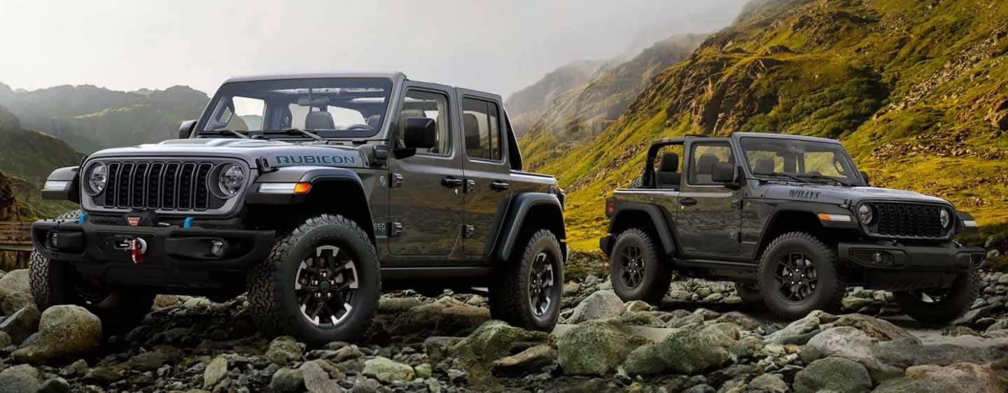 Dos modelos de Jeep Wrangler estacionados en un claro rocoso en las montañas. A la izquierda, un Wrangler 4xe Rubicon gris de cuatro puertas. A la derecha, un Wrangler Willys gris de dos puertas.