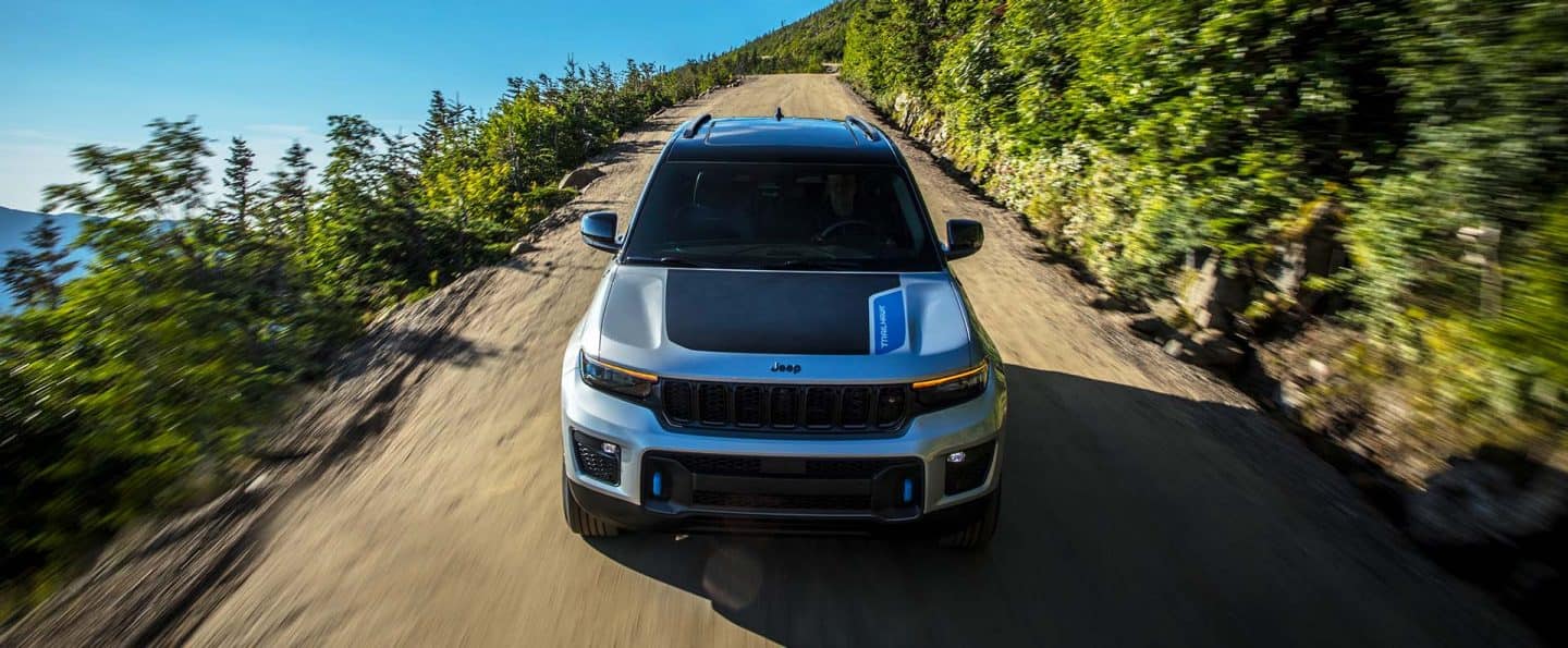 Ángulo frontal de un Jeep Grand Cherokee Trailhawk 4xe 2025 plateado con una inserción negra en el capó, circulando por un camino de tierra en las montañas, cerca de un cuerpo de agua.