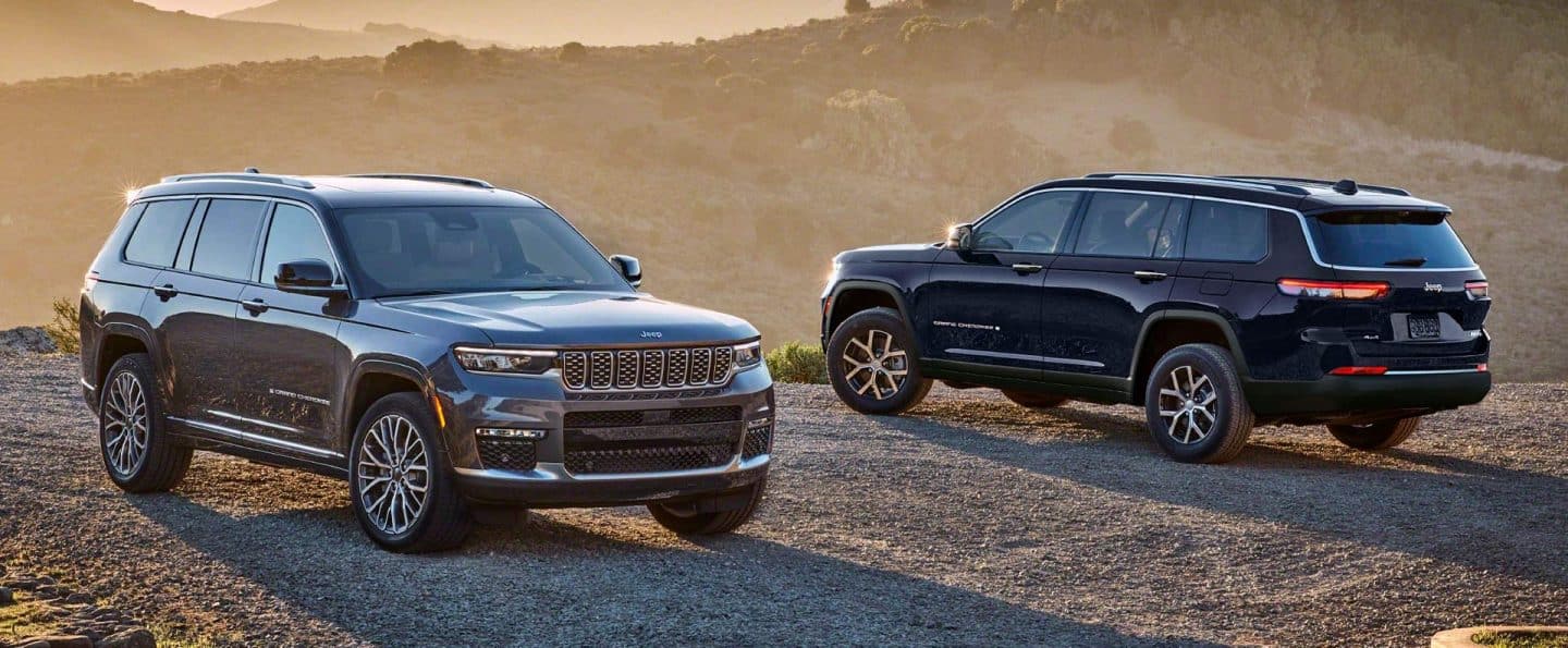 Un Jeep Grand Cherokee Summit Reserve 2025 gris y un Jeep Grand Cherokee Limited negro estacionados uno frente al otro en un camino de montaña de grava al atardecer.