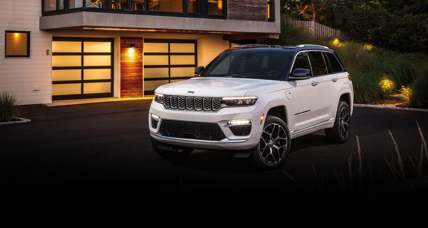 A white 2024 Jeep Grand Cherokee Summit Reserve 4xe with its headlamps on, parked in the driveway of a luxurious contemporary home at night.