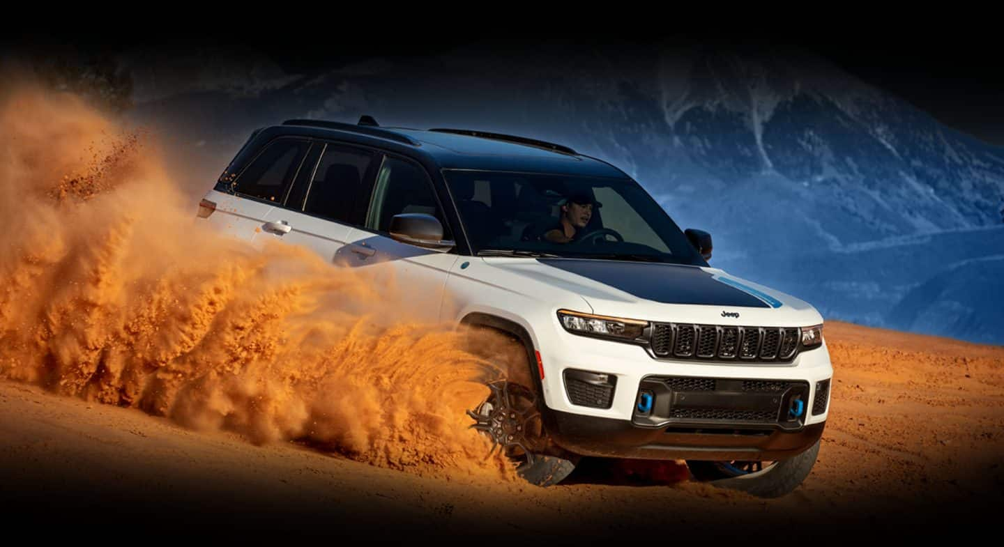 A white 2024 Jeep Grand Cherokee Trailhawk 4xe with a black hood insert, kicking up a cloud of orange sand and dust as it's being driven down a dune, with mountains in the background. 