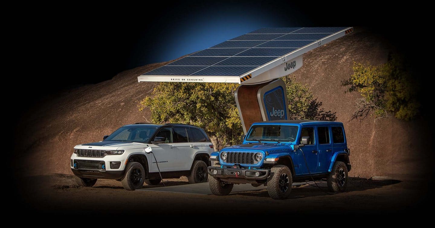 A white 2024 Jeep Grand Cherokee Trailhawk 4xe with a black hood insert, and a blue 2024 Jeep Wrangler Rubicon 4xe, both being charged at a Jeep-branded solar-powered charging station located off-road, with a huge hill in the background.