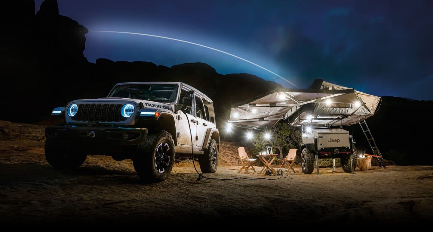 A 2024 Jeep Wrangler Rubicon 4xe parked beside a campsite at night, with one end of an inverter cord plugged into the vehicle's charging port and the other plugged into camping equipment. The campsite has a Jeep branded camper set up with awnings and lights.