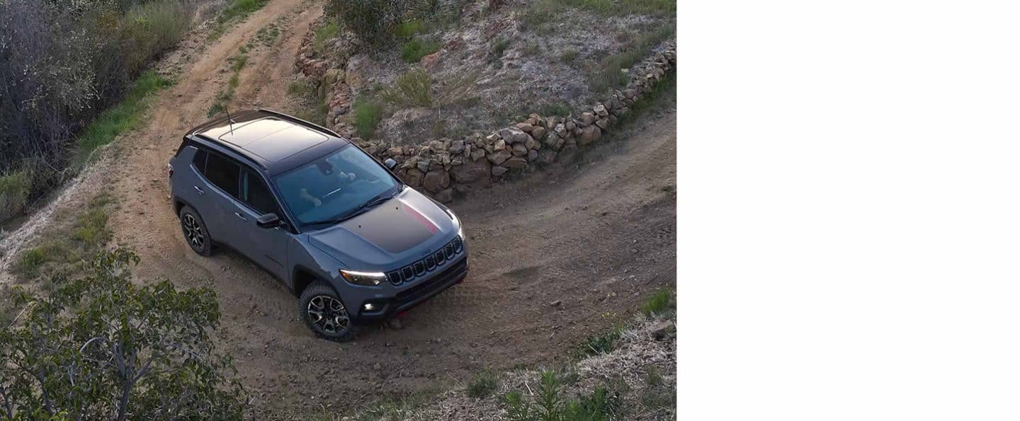 Un Jeep Compass Trailhawk 2024 circulando por la curva cerrada de un camino de tierra junto a un cerco de piedras en una colina.