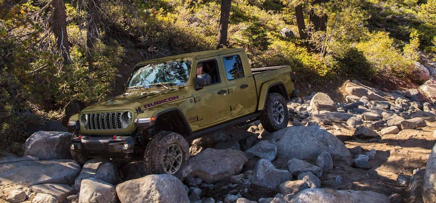 Una Jeep Gladiator Rubicon X 2025 verde oliva trepando por una serie de rocas en un bosque todoterreno.