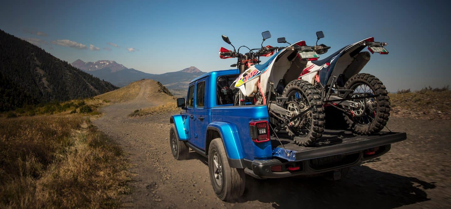 La parte posterior de un Jeep Gladiator Mojave X 2025 circulando por una carretera todoterreno con la puerta trasera abierta y dos vehículos todoterreno atados en la plataforma.