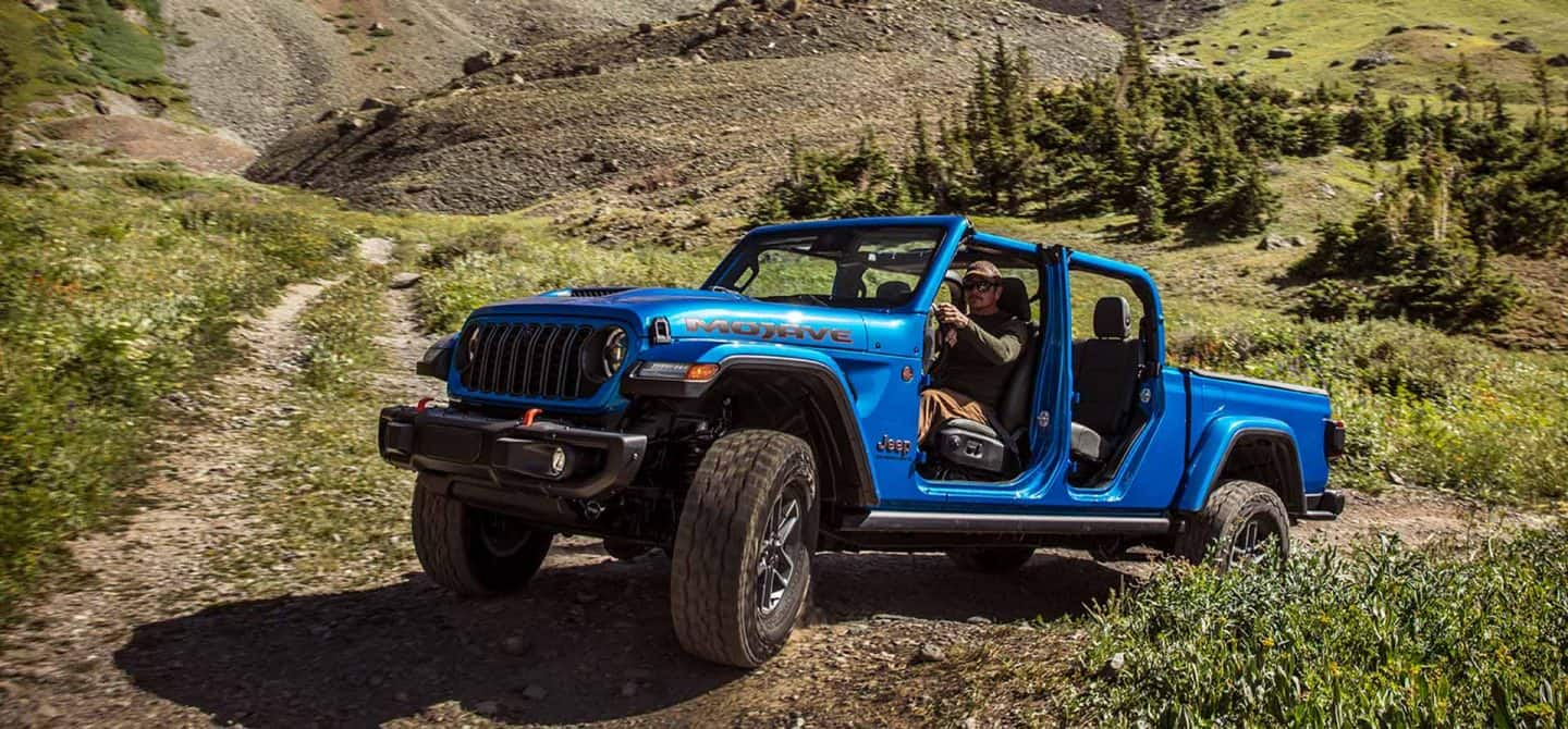 Una Jeep Gladiator Mojave X 2025 azul sin las puertas ni el techo, ascendiendo por un sendero en un área con colinas todoterreno.