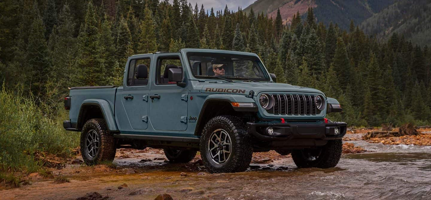 Una Jeep Gladiator Mojave X 2025 azul sin las puertas ni el techo, ascendiendo por un sendero en un área con colinas todoterreno.