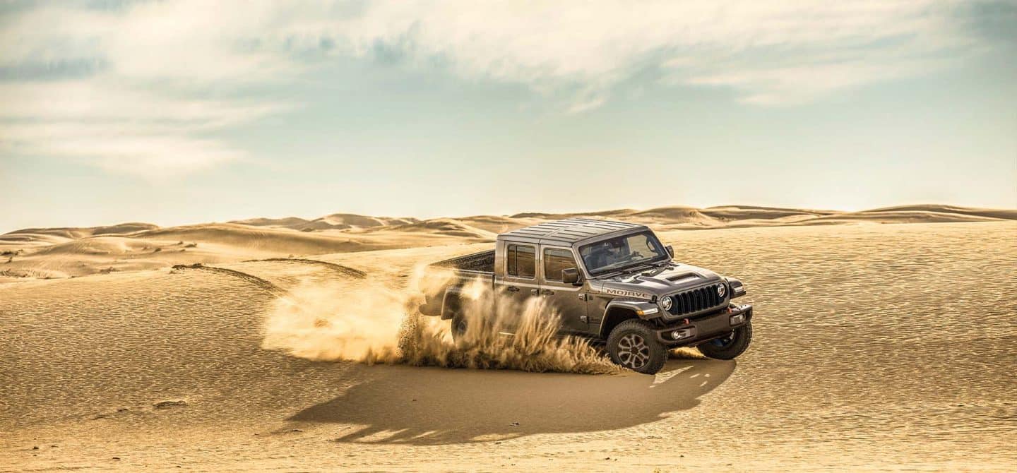 Una Jeep Gladiator Mojave X 2025 gris levantando arena mientras circula por un terreno desértico.