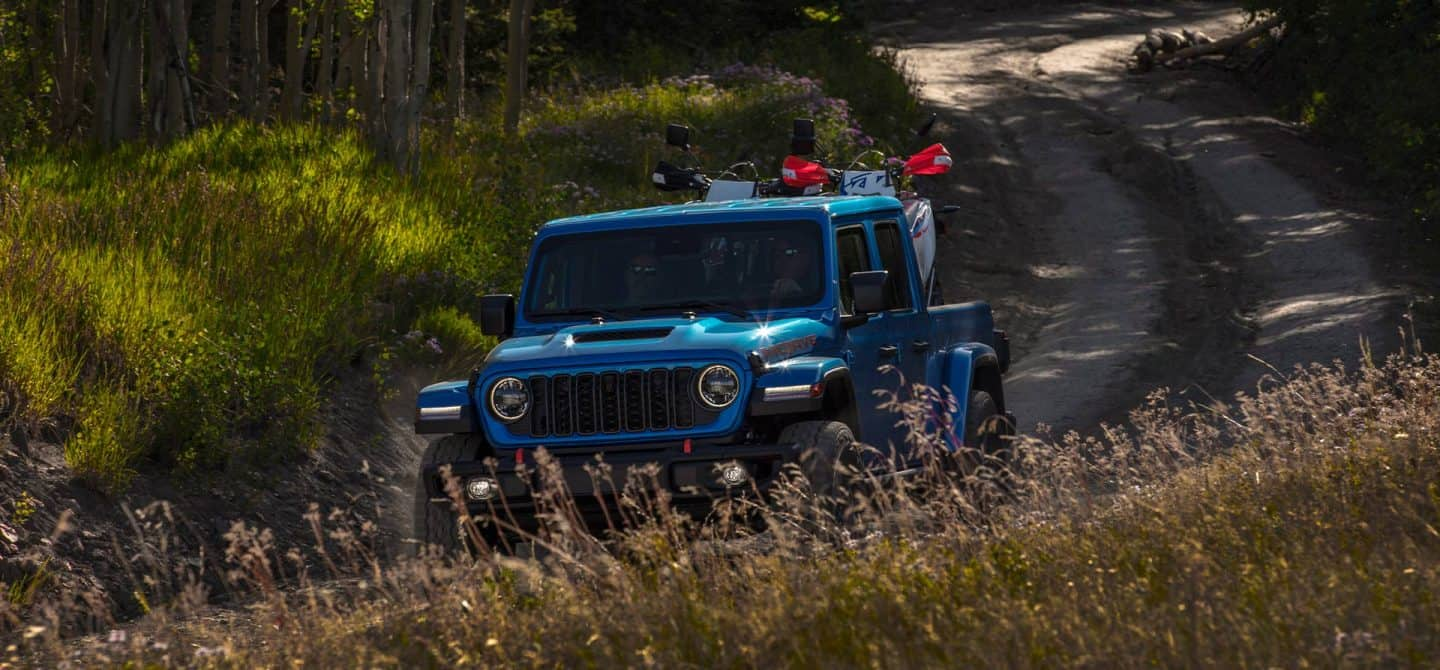 Una Jeep Gladiator Rubicon X 2025 azul circulando por una carretera rural sinuosa con dos vehículos todoterreno en la plataforma.