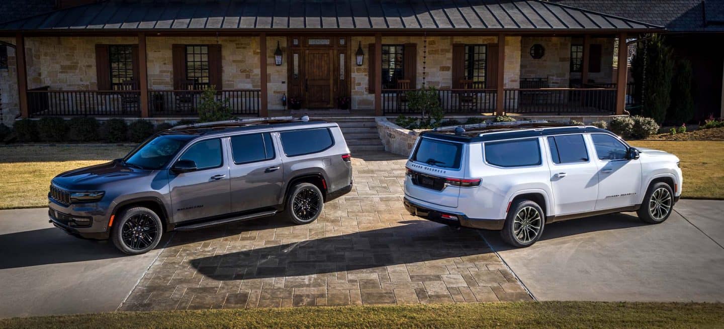 Dos modelos Jeep Grand Wagoneer estacionados uno frente al otro en el patio de un rústico edificio comercial. A la izquierda, un Jeep Grand Wagoneer Obsidian 2025 gris y a la derecha, un Jeep Grand Wagoneer Serie 3 2025 blanco.