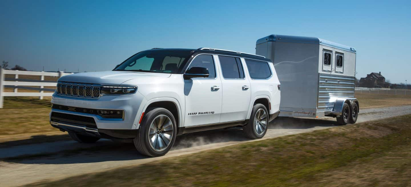 Un Jeep Grand Wagoneer Serie 3 blanco del año 2025 circulando por un camino de tierra al lado de una granja, remolcando un remolque para caballos.