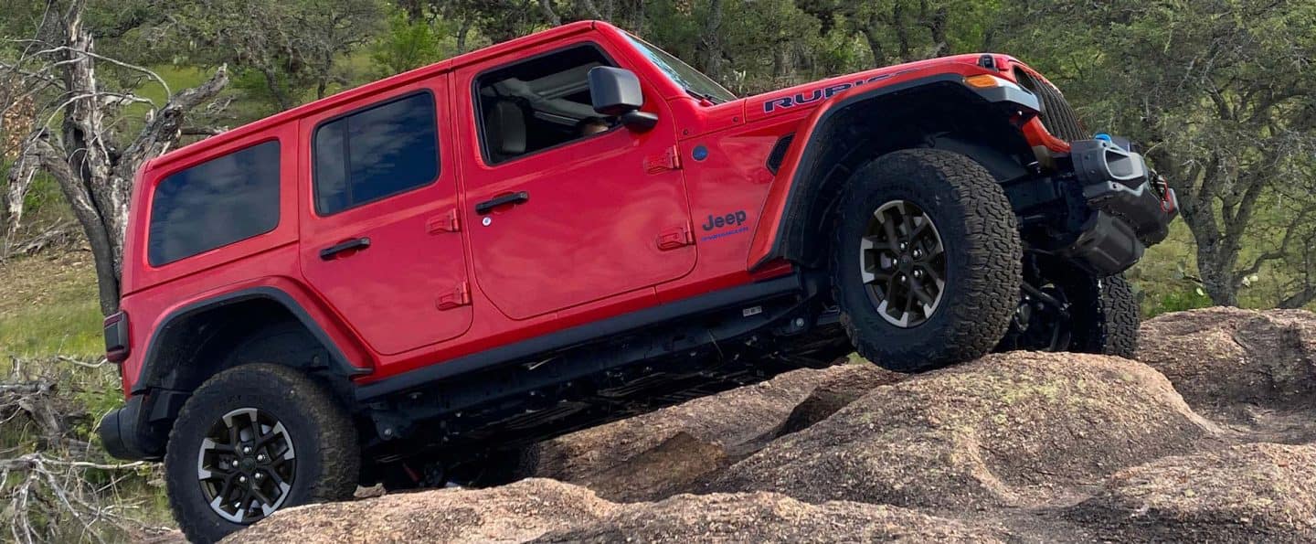 Perfil del lado del pasajero de un Jeep Wrangler Rubicon X 4xe 2025 rojo trepando una colina de peñascos cubiertos de arena.