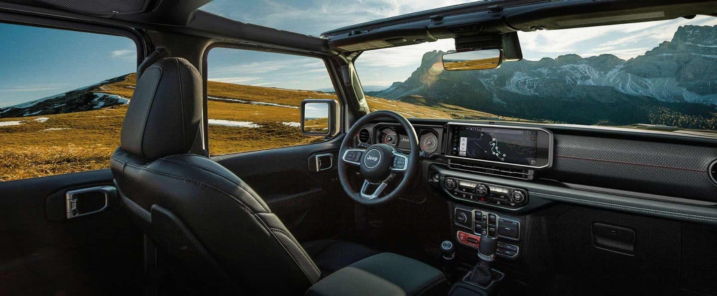 La fila delantera del Jeep Wrangler 2025 con el foco puesto en el techo corredizo abierto, el volante, la pantalla táctil de Uconnect y el tablero.