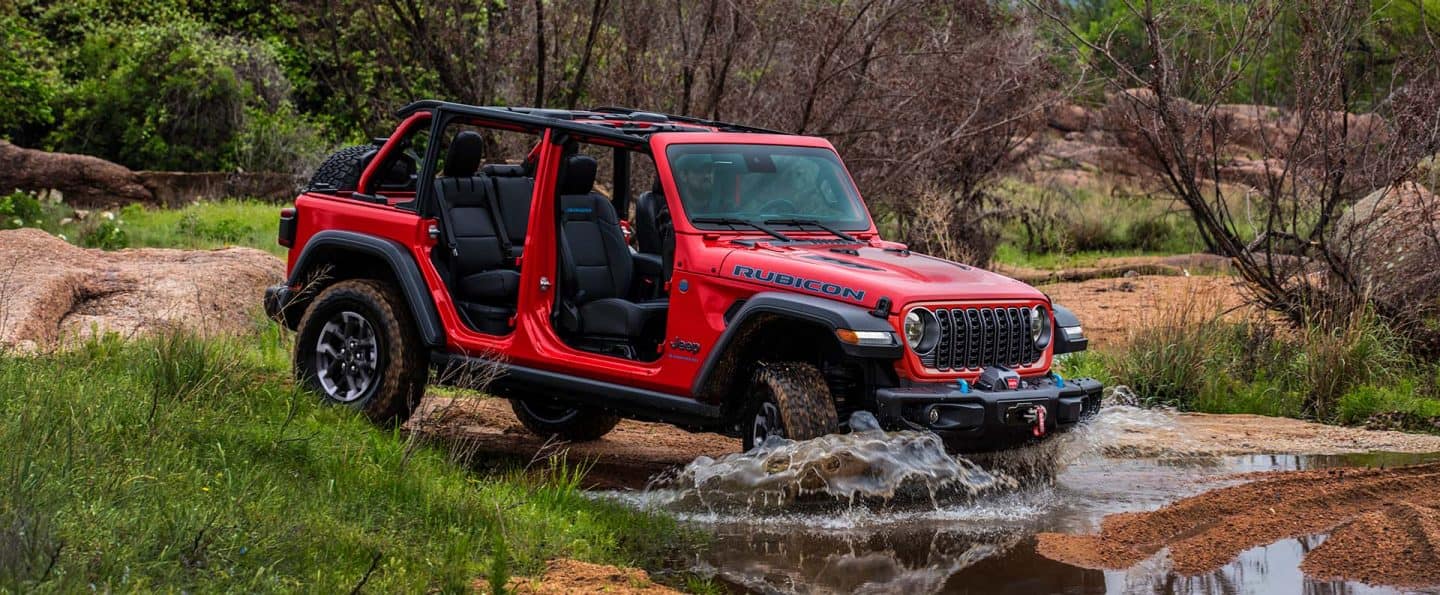 Un Jeep Wrangler Rubicon 4xe 2025 rojo con las puertas y el techo extraídos, ingresando a un arroyo, fuera de la carretera. 