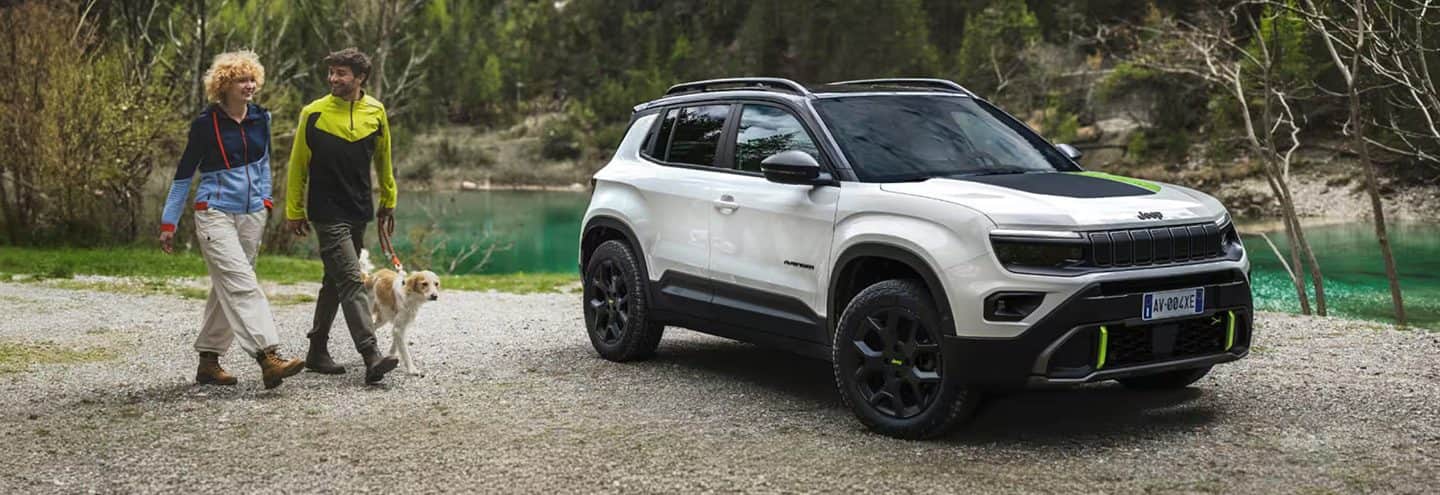 A white 2023 Jeep Wrangler Rubicon 4xe and a lime green 2023 Jeep Gladiator Rubicon, both with their doors and roofs removed, parked opposite of each other on sand at the foot of a large, rocky hill. Memorial Day Sales Event.