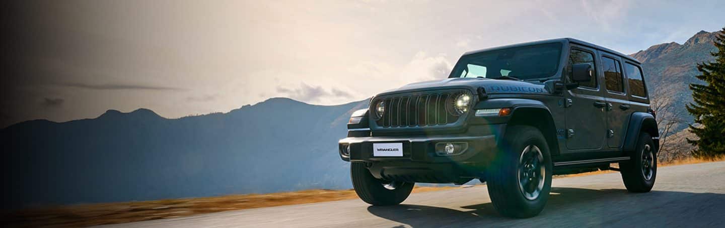 A white 2023 Jeep Wrangler Rubicon 4xe and a lime green 2023 Jeep Gladiator Rubicon, both with their doors and roofs removed, parked opposite of each other on sand at the foot of a large, rocky hill. Memorial Day Sales Event.