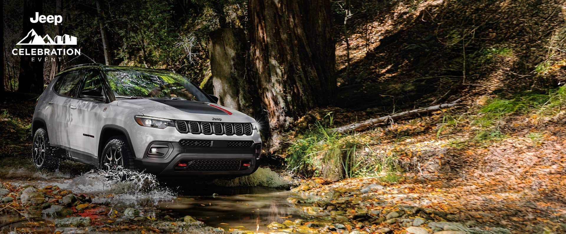 A white 2024 Jeep Compass Trailhawk with a black hood insert, fording through a shallow stream off-road in the woods. Jeep Celebration Event.