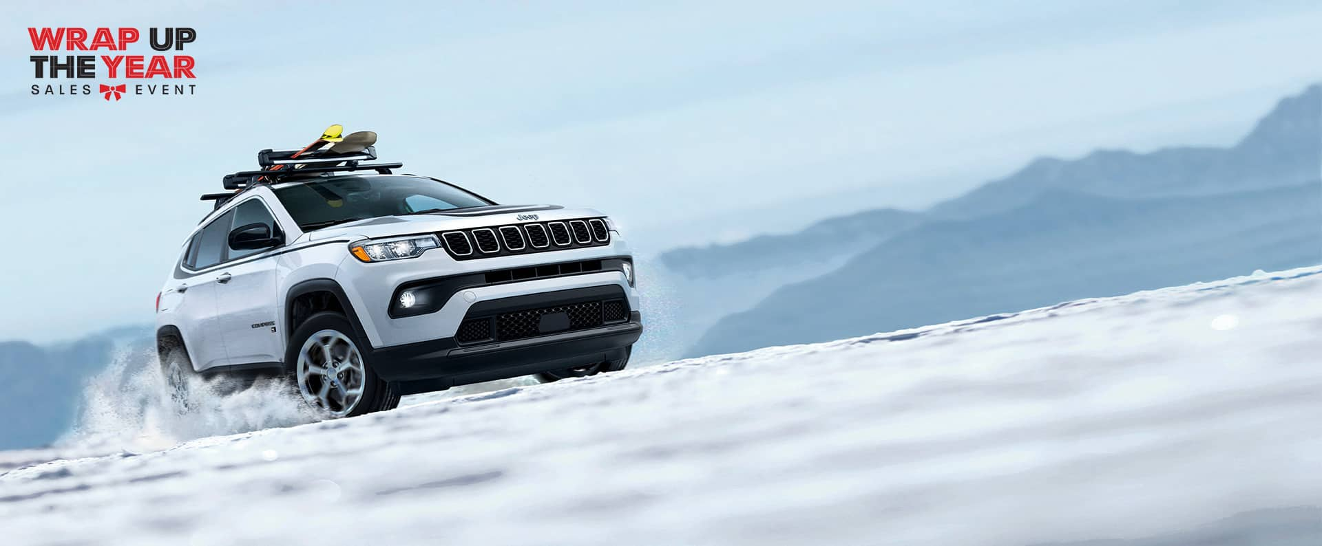 A white 2024 Jeep Compass Latitude with ski equipment secured on its roof rack, ascending a snow-covered hill off-road. Wrap Up The Year Sales Event logo.