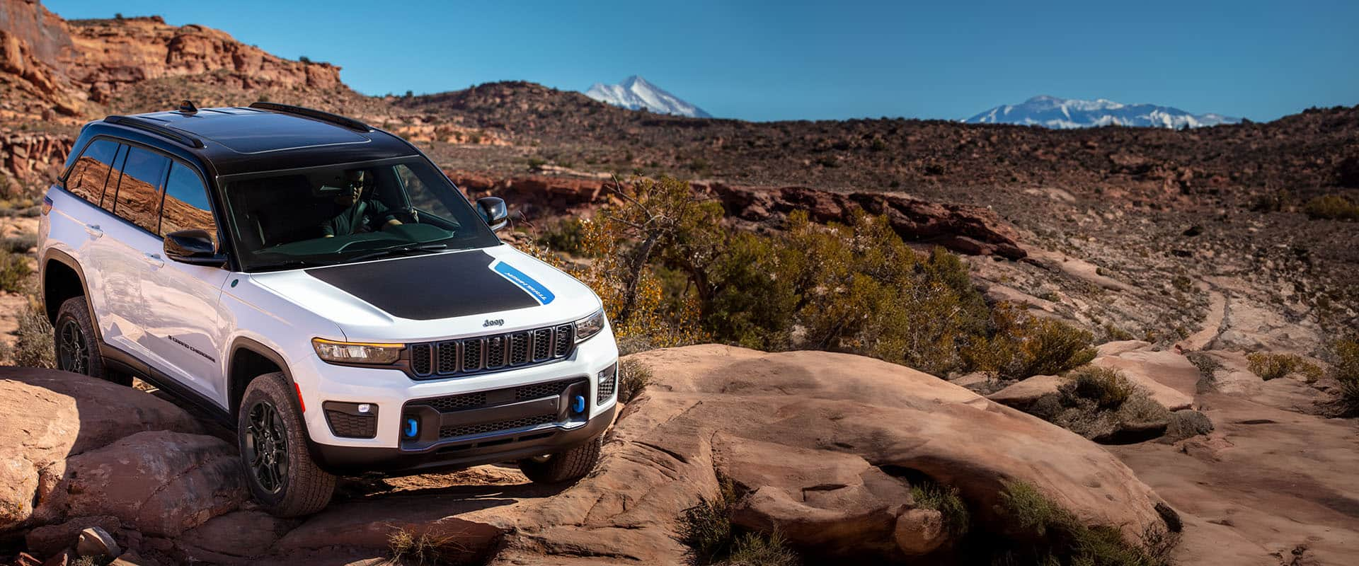 A white 2024 Jeep Grand Cherokee 4xe Trailhawk crawling over large boulders in the desert, with mountains in the background.