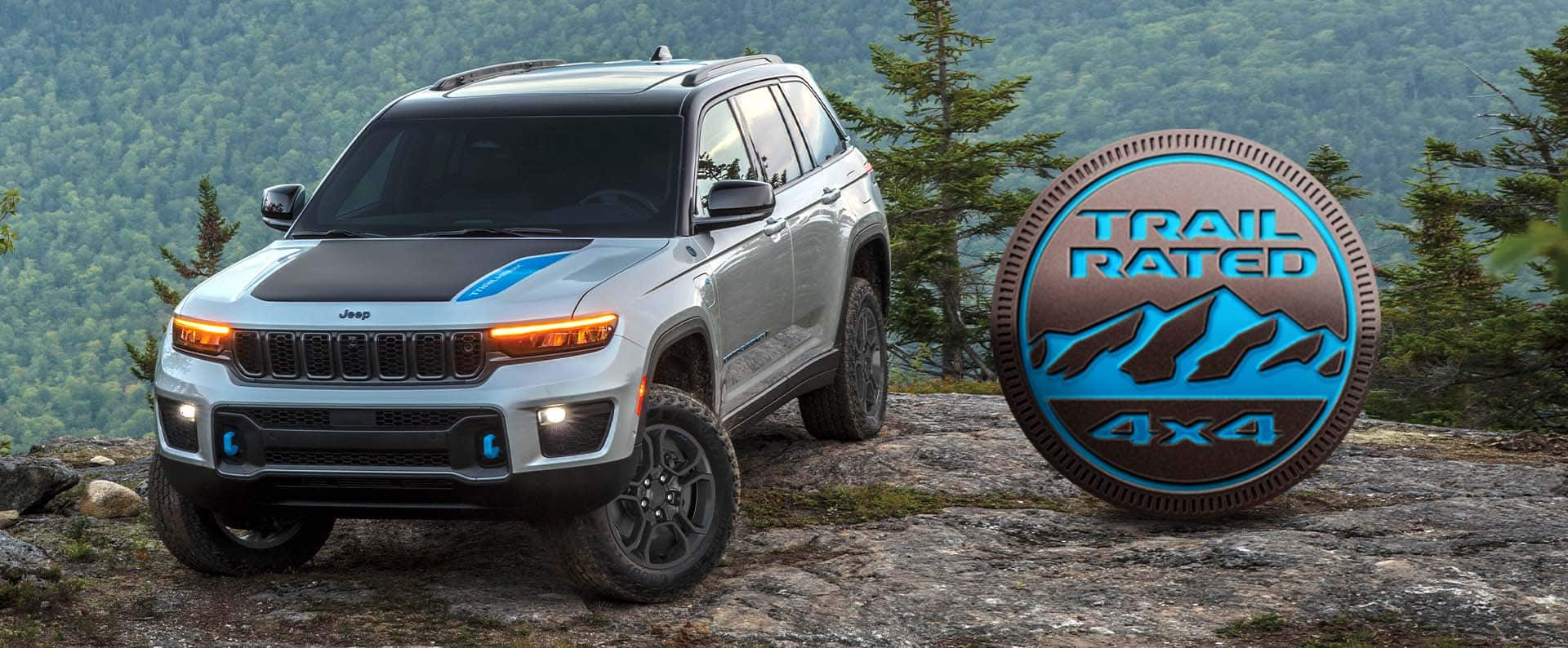 A silver 2024 Jeep Grand Cherokee Trailhawk 4xe parked on a clearing off-road, with a mountain forest in the background. The Trail Rated 4x4 badge.