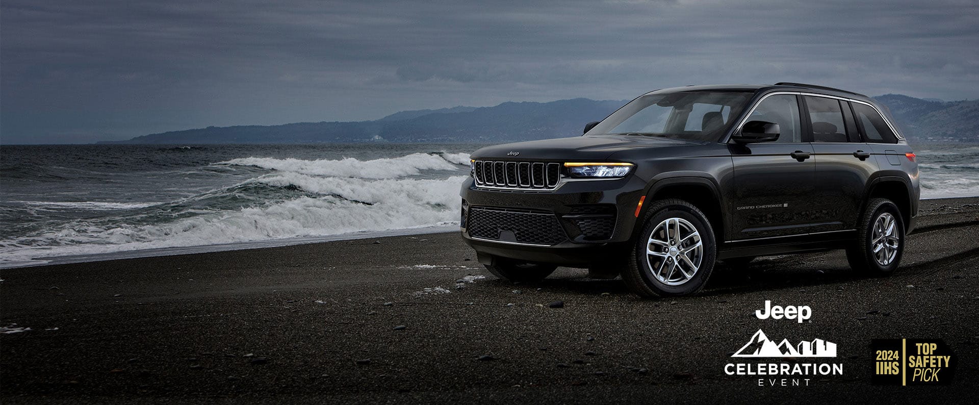 A black 2024 Jeep Grand Cherokee Laredo parked on a beach at the water's edge with storm clouds overhead. Jeep Celebration Event.