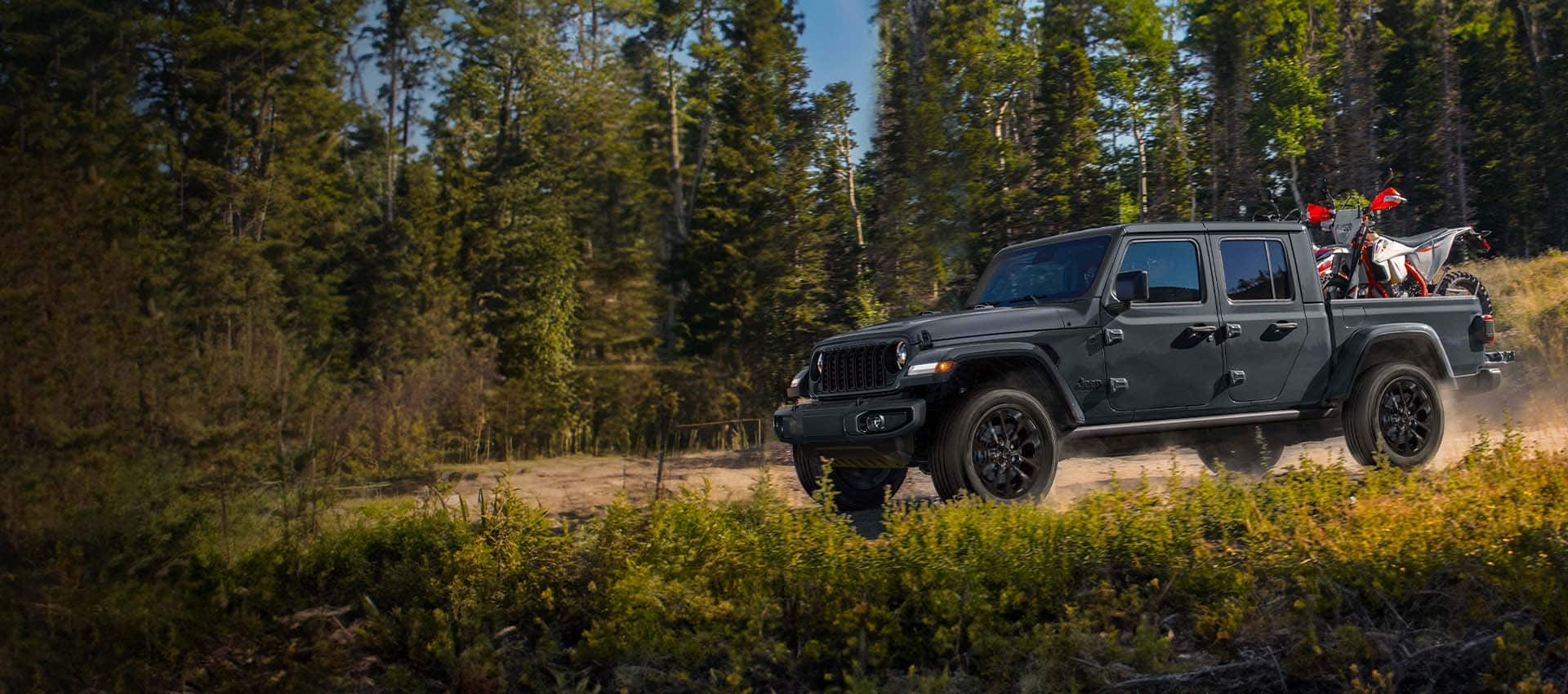 Un Jeep Gladiator Nighthawk 2024 circulando por un sendero todoterreno en el bosque con un ATV en su plataforma.