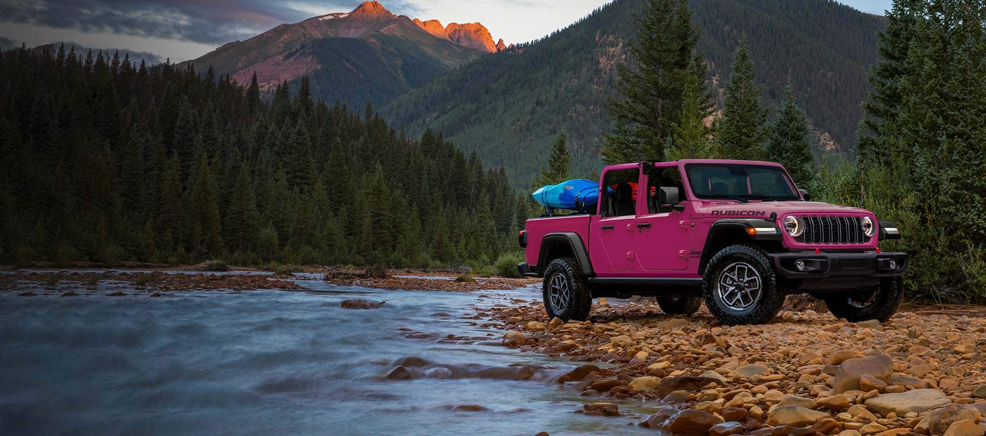 Vista en ángulo delantera del lado del pasajero de un Jeep Gladiator Rubicon 2024 estacionado sobre un lecho de rocas al costado de un arroyo, con equipos en la caja de la camioneta y un bosque de montaña denso de fondo.