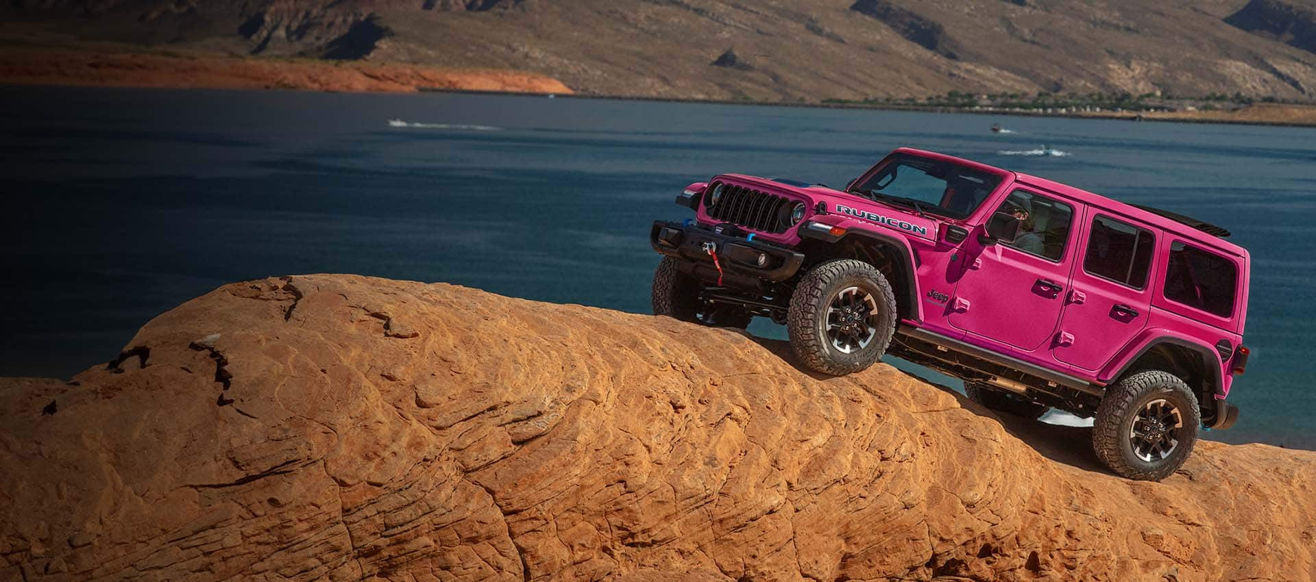 A hot pink 2024 Jeep Wrangler Rubicon four-door ascending a steep, rock hill off-road, with a body of water in the background.