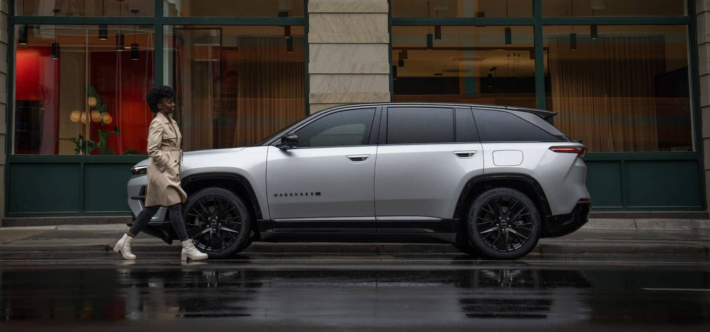 Display A driver-side profile of a silver 2024 Jeep Wagoneer S Launch Edition parked on a rain-covered street in front of an upscale business in a metropolitan neighborhood.