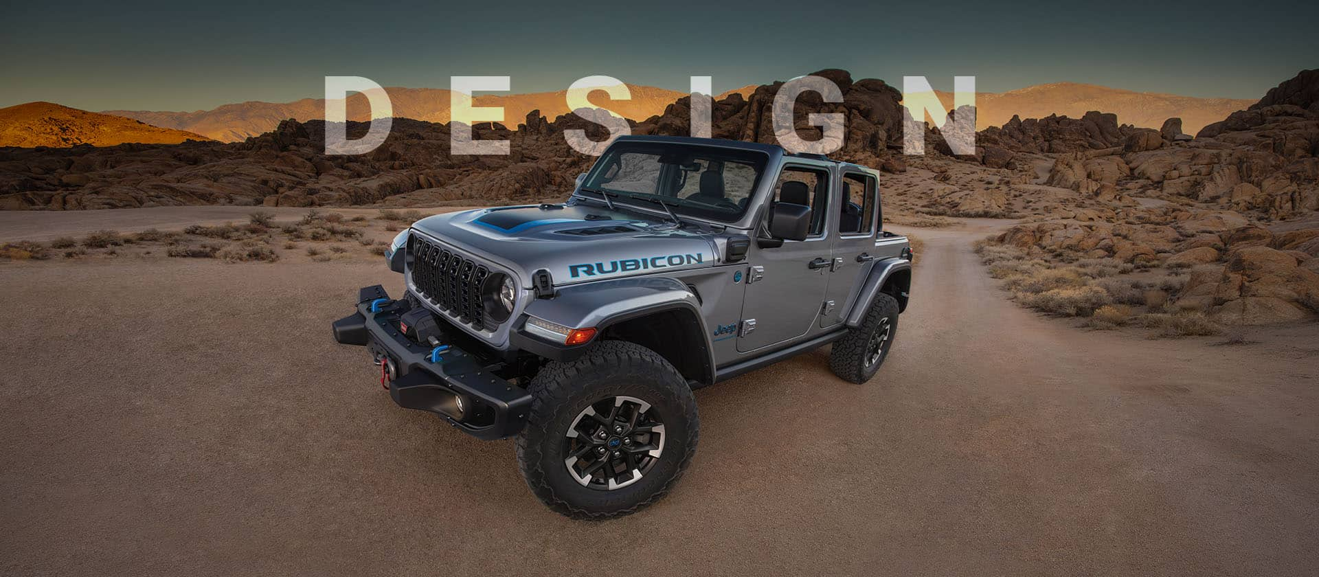 An angled driver-side profile of a gray 2024 Jeep Wrangler Rubicon 4xe parked on a clearing in the desert. Design.