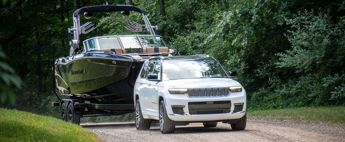 A white 2025 Jeep Grand Cherokee Summit Reserve traveling down a dirt road towing a large motorboat.