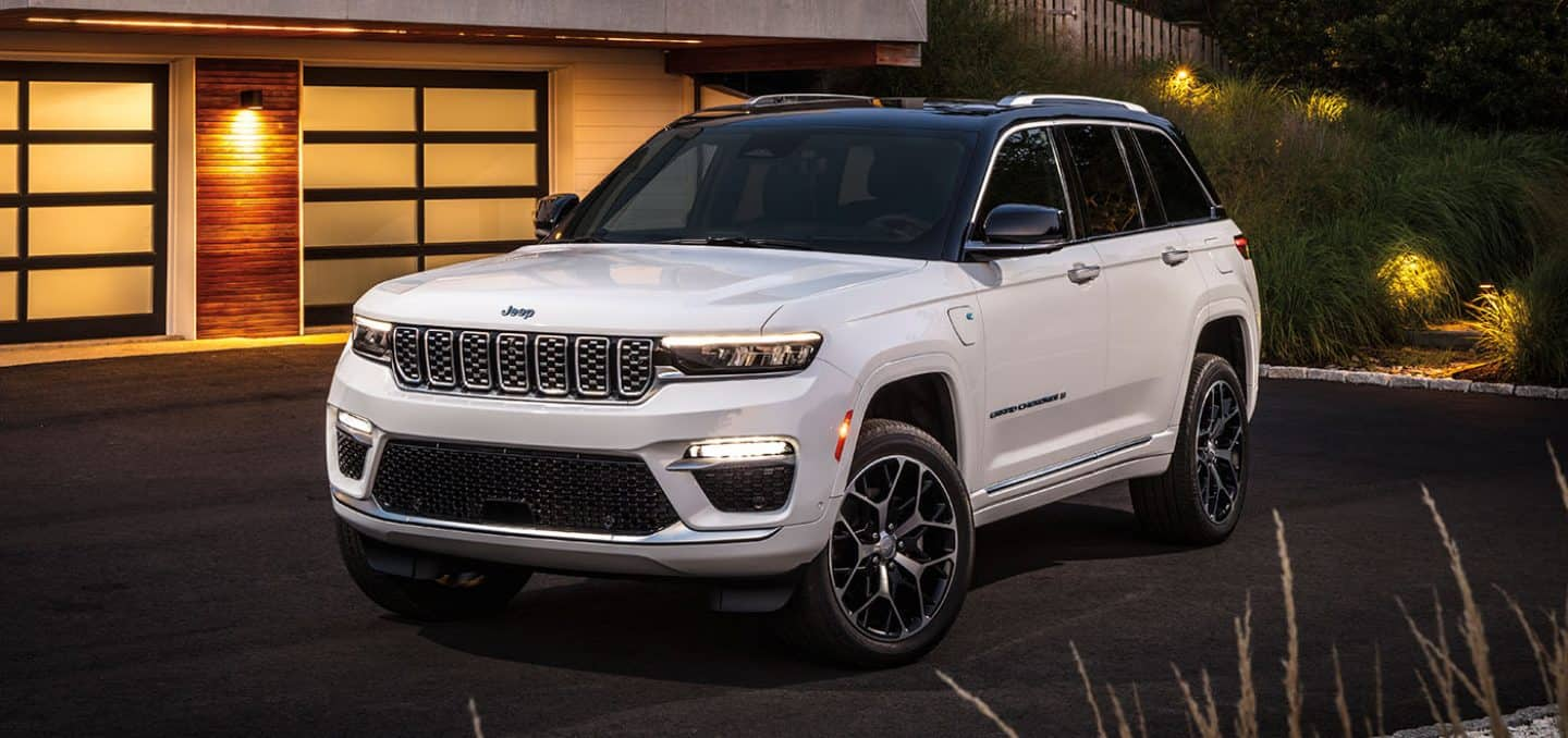 Display A white 2025 Jeep Grand Cherokee Summit Reserve 4xe parked outside of a contemporary home in the evening. 