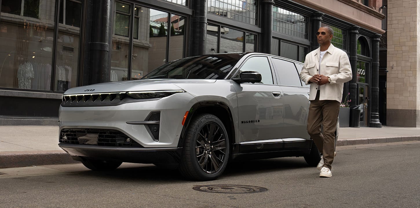 A silver 2025 Jeep Wagoneer S Launch Edition parked on a street in a metropolitan neighborhood with a man walking nearby.