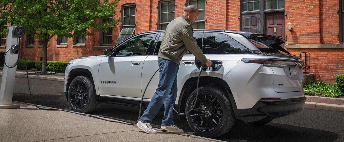 A silver 2025 Jeep Wagoneer S Launch Edition with a man plugging the charging cord from a public charging station into the vehicle's charging port.